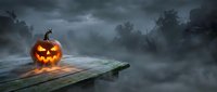 Spooky Halloween jack-o-lantern pumpkin glowing on wooden dock in misty swamp with dramatic stormy sky and bare trees creating eerie atmospheric scene.