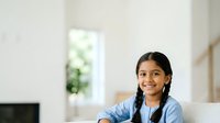 Young South Asian woman with long dark braided hair wearing light blue denim shirt, smiling at camera in bright modern interior space, suitable for lifestyle and portrait projects.