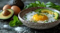 Sunny side up egg in rustic ceramic bowl with fresh avocado slices, herbs and coarse salt on dark background, healthy breakfast composition.