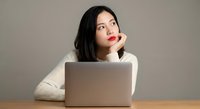 Young Asian woman in white sweater sitting at wooden desk with laptop computer, looking thoughtful while resting chin on hand in modern workspace.