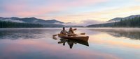 Two people fishing from wooden boat on calm lake at sunrise with forested mountains and pink sky reflections, outdoor recreation and nature scene.