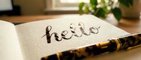 Fountain pen resting on open notebook with handwritten hello in elegant calligraphy, warm natural light, green plant in background.