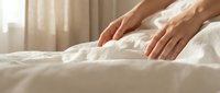 Close-up of hands touching soft white bedding in bright morning light, suitable for hospitality, wellness, and lifestyle marketing materials.