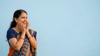 Young South Asian woman wearing traditional orange and blue patterned kurta, expressing joy and surprise with hands near face against light blue background.