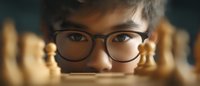 Young Asian male in glasses peering through chess pieces on board, focused gaze behind black frames, strategic perspective in warm lighting atmosphere.