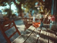 Glass of rosé wine on wooden table at outdoor restaurant terrace during golden hour with warm bokeh lights creating romantic dining atmosphere.