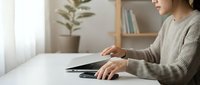 Young Asian woman in beige sweater using smartphone at white desk in modern home office with plant and bookshelf background for remote work lifestyle.