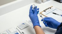 Healthcare worker in blue gloves preparing vaccine injection in medical clinic setting with forms and supplies on table.
