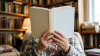 Senior woman reading open book in cozy home library with wooden shelves and warm lighting, suitable for education, literacy, and lifestyle content.
