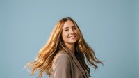 Happy young Caucasian woman with flowing blonde hair in motion wearing beige coat against blue sky background expressing joy and freedom in lifestyle portrait.