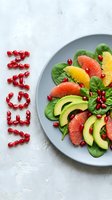 Vegan salad with spinach, avocado, grapefruit, orange, and pomegranate seeds spelling vegan on a light concrete background.