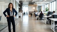 Confident female professional with dark hair and Mediterranean ethnicity wearing black blazer standing in modern office corridor with colleagues working in background.