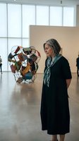 Senior Caucasian woman with gray hair wearing black dress and green scarf viewing contemporary art sculpture in modern gallery with large windows.