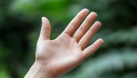 Open palm of a Caucasian person reaching up against blurred green natural background, showing five fingers in welcoming or spiritual gesture. Soft focus creates peaceful mood.