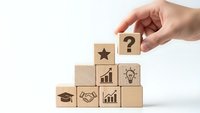 Hand placing wooden cube with question mark on pyramid of blocks featuring business icons including star, chart, lightbulb, handshake and graduation cap.