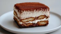 Homemade tiramisu dessert with layers of coffee-soaked ladyfingers, mascarpone cream, and cocoa powder dusting on white plate.