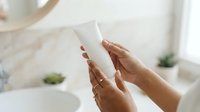 Young woman with medium skin tone holding blank white cosmetic tube in bright bathroom setting for skincare product mockup.