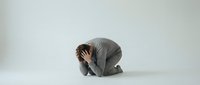 Businessman in gray suit kneeling with head in hands showing despair and stress on white background for mental health and business failure concepts.