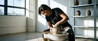 Young Asian male potter shaping clay on spinning wheel in bright modern studio with pottery displayed on shelves. Artisan craftsmanship and creative process.