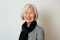 Elegant senior Asian woman with gray hair and bright smile wearing black scarf and gray blazer against light background, representing active aging and wellness.
