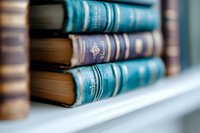 Vintage hardcover books with ornate spines in shades of blue and turquoise arranged on white wooden shelf, shallow depth of field creates dreamy bokeh effect.