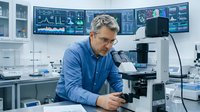 Middle-aged Caucasian male scientist in blue shirt working with microscope in modern laboratory with multiple computer monitors displaying data and research.