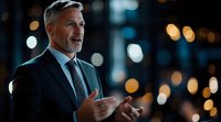 Mature Caucasian businessman with gray hair and beard gesturing while speaking, wearing dark suit against blurred city lights background in evening setting.