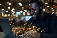 Young African American male customer service representative wearing headset and glasses works on laptop with magical glowing network effect, smiling at night office.