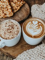 Two coffee cups with latte art and foam designs served on wooden board next to pastries, cozy cafe setting with knitted textile, warm moody atmosphere.