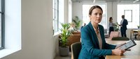 Professional businesswoman in teal blazer working on tablet in modern office space with colleagues in background during daytime meeting.