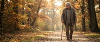 Senior Caucasian man with walking stick strolling through autumn park path surrounded by golden fall foliage and warm sunlight filtering through trees.