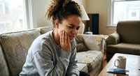 Young African American woman experiencing tooth pain sitting on couch at home holding jaw with hand showing dental discomfort and oral health issues.