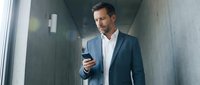 Middle-aged man with dark hair and facial stubble, appearing to be of Mediterranean or Latin descent, wearing a dark blazer and white shirt, holding a smartphone while walking through a modern concrete corridor or hallway.