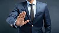 Businessman in navy blue suit showing stop gesture with palm forward against dark background, conveying authority and boundary setting.