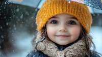 Young child wearing yellow knit beanie and beige scarf smiling during snowfall, close up portrait with bright blue eyes and joyful expression in winter weather.