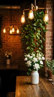 Cozy industrial interior with vintage Edison bulb chandelier, white flowers in ceramic vase on wooden table, green ivy plant against exposed brick wall.