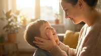 Young Caucasian mother tenderly holding her toddler son's face while both smile joyfully in warm sunlit home interior, expressing pure maternal love and connection.