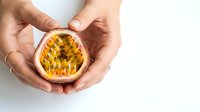 Hands holding halved passion fruit with visible seeds and pulp against white background, suitable for food and culinary design projects.