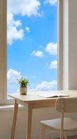 Wooden desk with open notebook and potted plant near large window overlooking blue sky with white clouds, bright natural light workspace for home office or study.