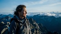 Middle-aged man with long dark hair and facial hair, wearing dark winter jacket, smiling while overlooking vast mountain range with snow-capped peaks under blue sky. Adventure and outdoor lifestyle concept.