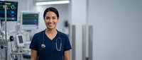 Smiling female healthcare professional in navy scrubs with stethoscope standing in modern hospital room with medical equipment and monitors.
