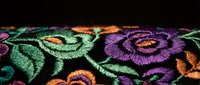 Colorful floral embroidery with roses in purple, green and orange threads on black fabric showcasing traditional needlework craftsmanship for textile design.