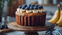 Fresh banana bread cake topped with sliced bananas and ripe blueberries on wooden cake stand, rustic kitchen setting with natural lighting and soft focus.