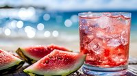Refreshing watermelon juice with ice cubes and fresh fruit slices on beach table with blurred ocean background. Summer tropical drink concept for vacation relaxation.