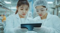 Asian female scientists in protective workwear analyzing data on tablet in modern laboratory setting, wearing hairnets and safety glasses while conducting research.