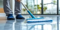 Professional cleaner mopping shiny floor surface in modern building interior, wearing blue work shoes and gray uniform pants, using blue microfiber mop.