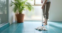 Woman mopping blue tile floor in bright modern home interior with palm plant by window. Household cleaning and maintenance concept for domestic lifestyle.