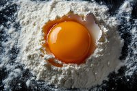 Fresh raw egg yolk in white flour on dark surface, macro food photography with dramatic lighting for cooking and baking concepts.