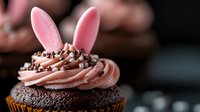 Chocolate cupcake with pink frosting topped with sprinkles and bunny ears decoration against dark background, selective focus on swirled buttercream topping.
