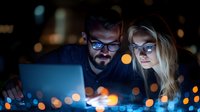 Young Caucasian couple working late on laptop with blue light reflection from screen, dark room with bokeh lights background, modern workplace concept.